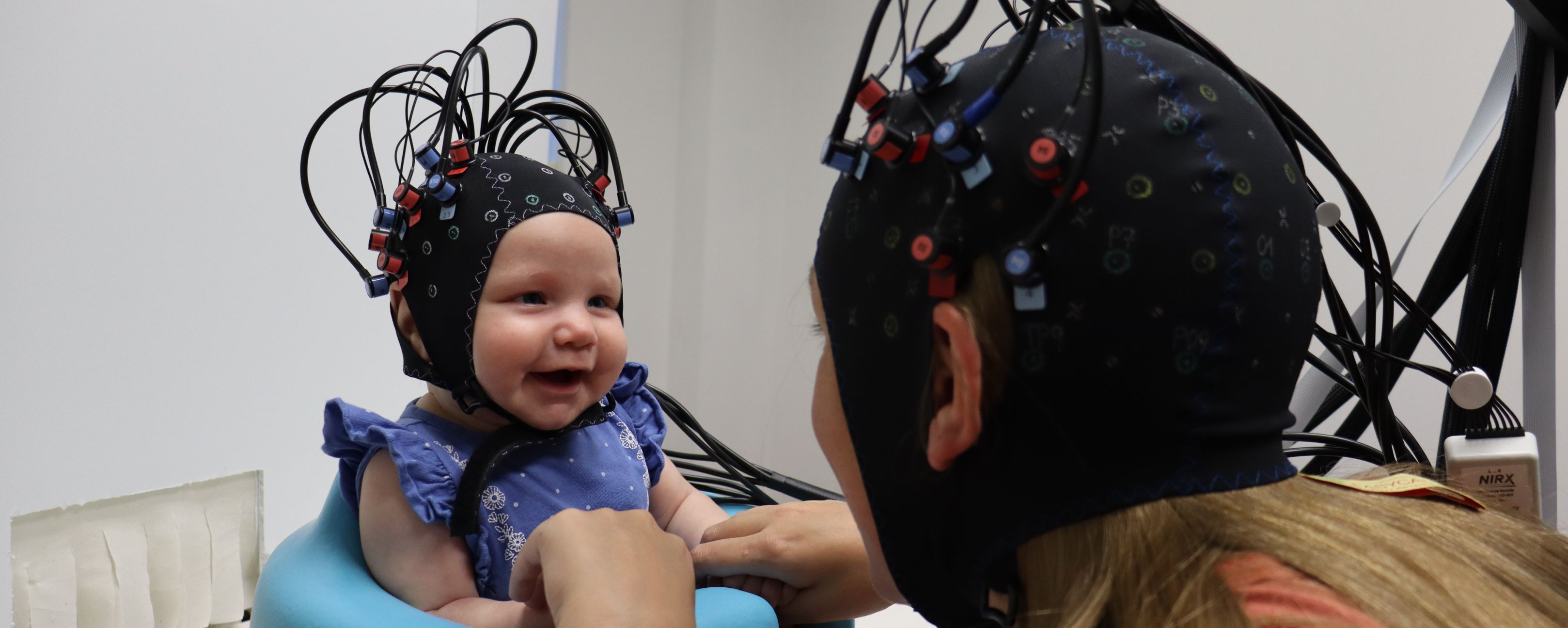 Infant and mother in neuroimaging headgear. Infant is smiling.